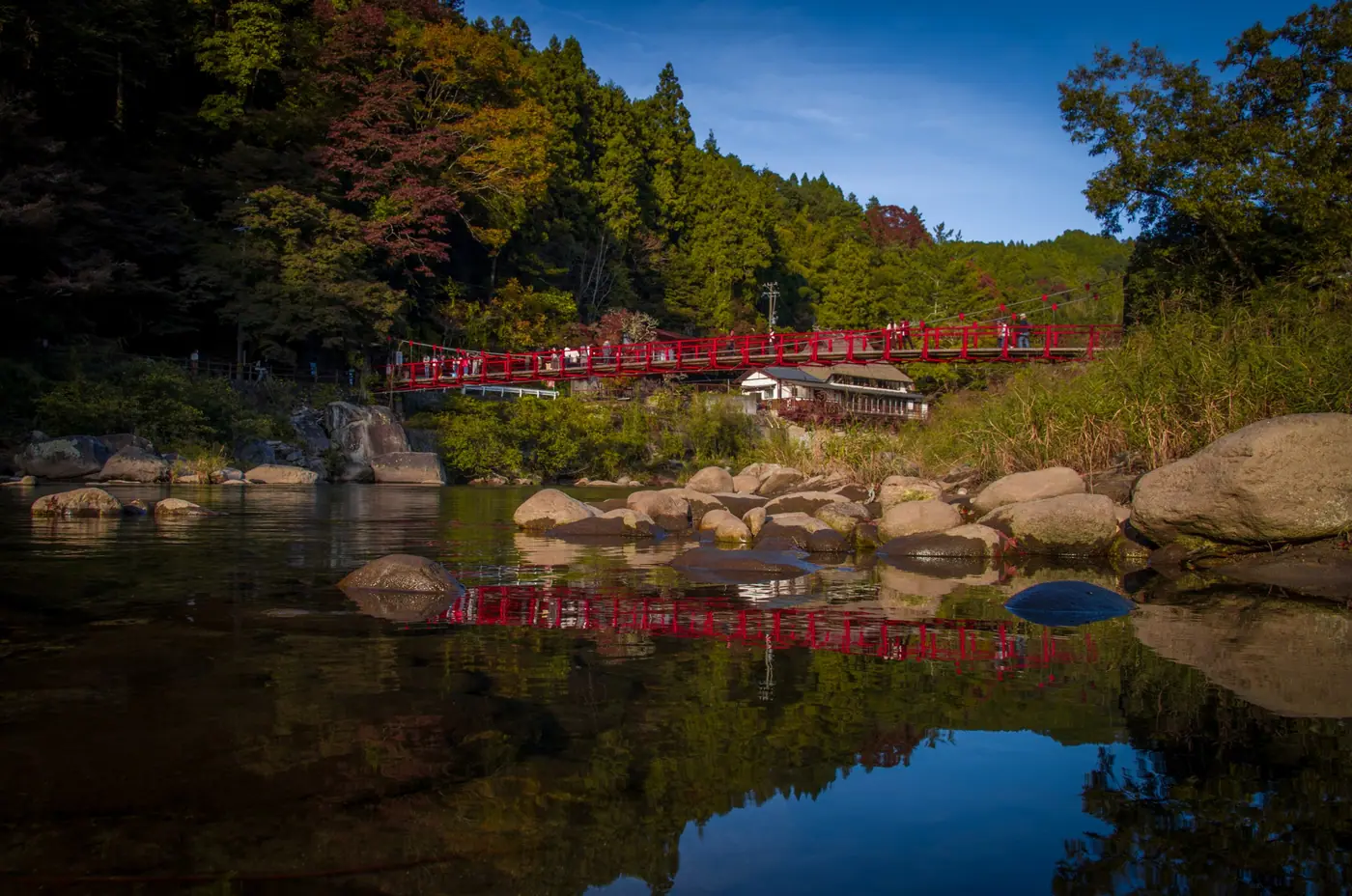 Rocks and still water below the red bridge, with trees just starting to turn