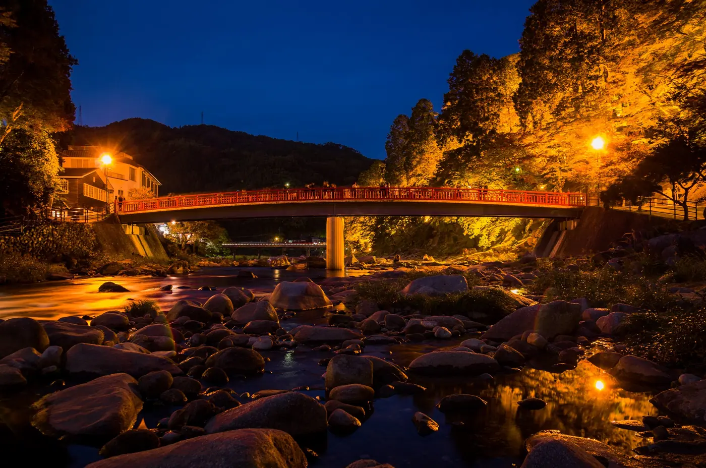 The Taigetsukyo Bridge lit up at dusk, with lanterns on the riverbank